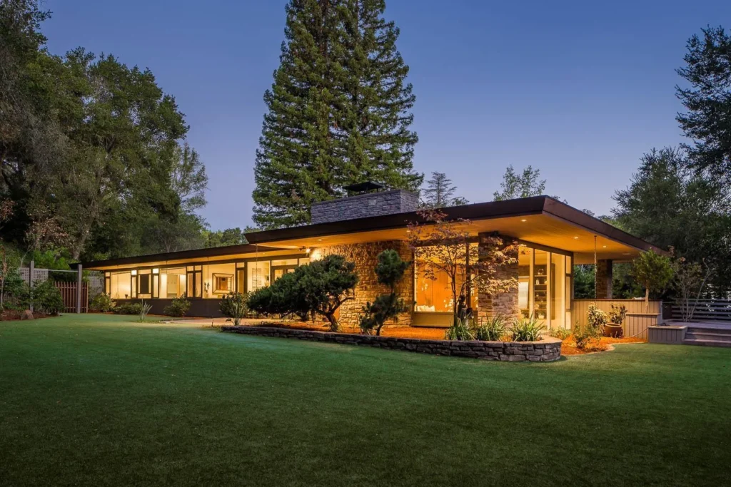 Mid-century modern home with large windows, stone accents, and landscaped yard at twilight