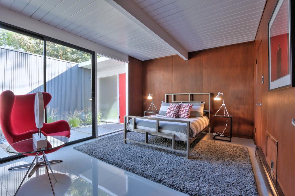 Mid-century modern Eichler bedroom with wood panel walls, red lounge chair, and floor-to-ceiling windows
